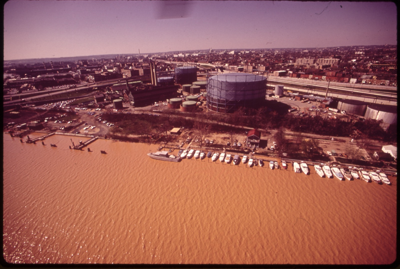 A 1973 color aerial of the Eastern Power Boat Club, its boats lined along the docks on a rust-orange, polluted Anacostia River, with riverside gasworks tanks and the city beyond.